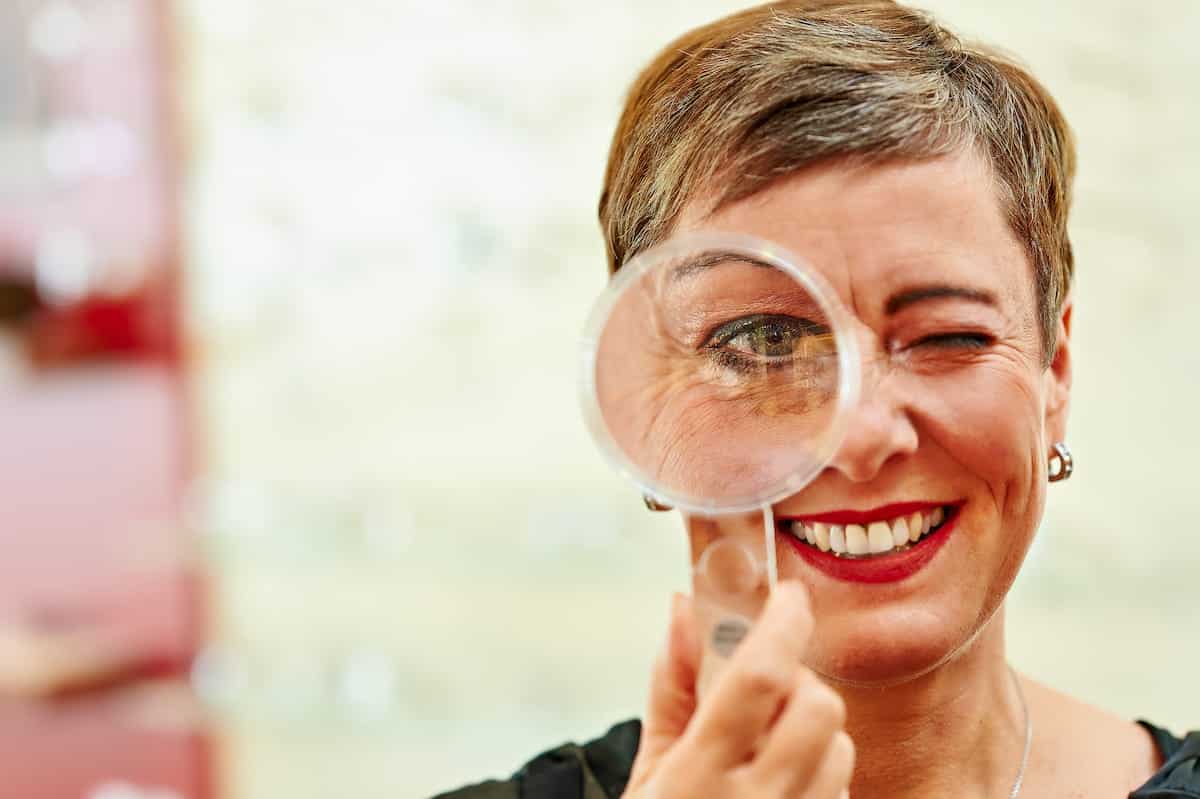 Frau mit Lupe roten Lippen und kurzen Haaren bei Optik Boysen, Frau mit Lupe in der Hand vor dem linken Auge
